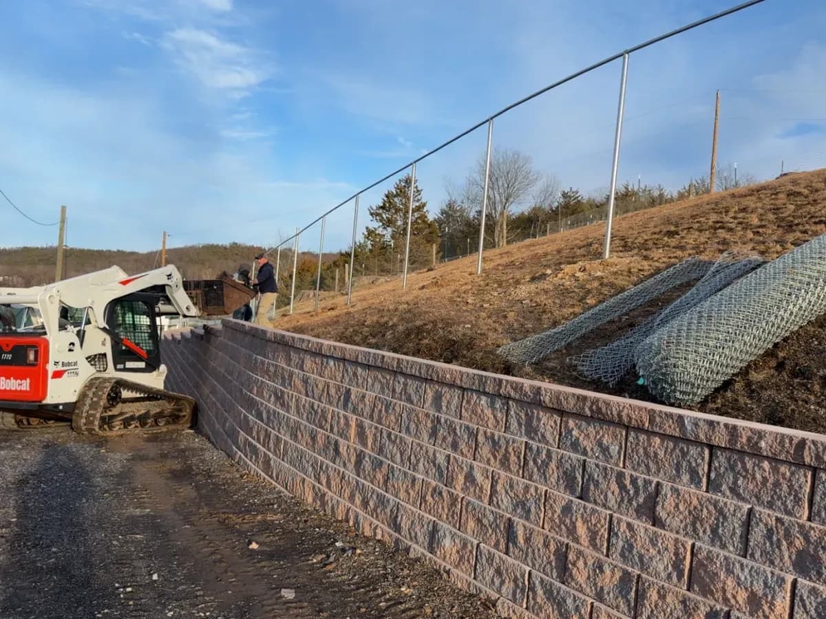 Compact track loader staged on a retaining wall during a chain-link install